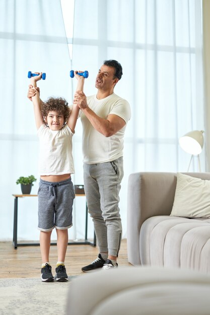 Volledige opname van een sportieve latijnse vader van middelbare leeftijd die zijn zoon leert oefenen met halters terwijl hij thuis tijd samen doorbrengt. actieve levensstijl, ouderschapsconcept