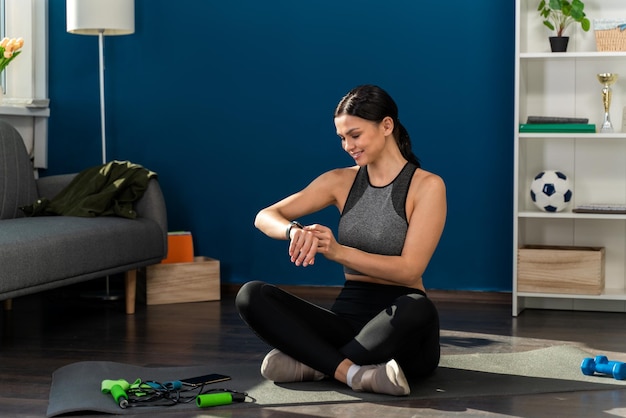 Volledige weergave van een glimlachende jonge vrouw die naar haar smartwatch kijkt terwijl ze thuis op de yogamat zit na de training