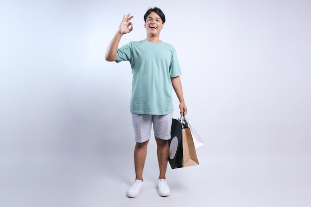 Volledige weergave van een jonge Aziatische man die casual zomerkleding draagt en een ok-bordje toont en winkelzakken vasthoudt