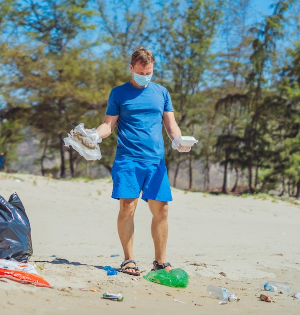 青いフェイスマスクのボランティア森の砂浜アクティブな男性がゴミを拾うプラスチック使い捨て食器バッグこぼれたゴミゴミ惑星汚染環境保護自然社会問題