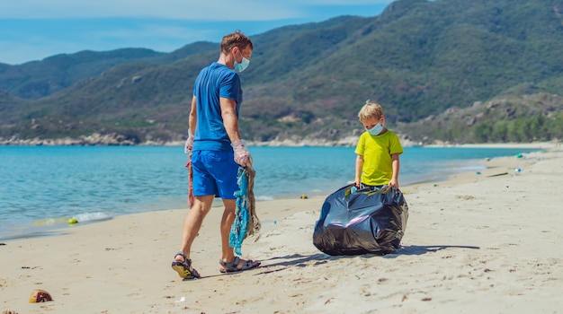 Volontario maschera viso blu lazur mare sabbia spiaggia il figlio aiuta il padre a tenere la borsa nera per raccogliere i rifiuti problema spazzatura versata spazzatura pianeta inquinamento protezione ambientale educazione naturale dei bambini