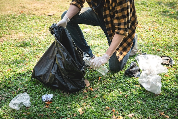 写真 ペットボトルとプラスチック廃棄物をプラスチックの黒い袋に拾い上げてゴミを収集する環境活動中に公園を掃除するために座っている手袋をはめたボランティアの男