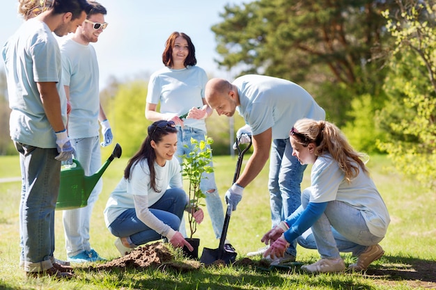 volunteering, charity, people and ecology concept - group of happy volunteers planting tree and digging hole with shovel in park