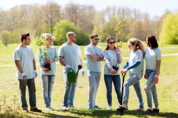 ボランティア、チャリティー、人々、エコロジーのコンセプト – 公園で木の苗やガーデニングツールを持つ幸せなボランティアのグループ