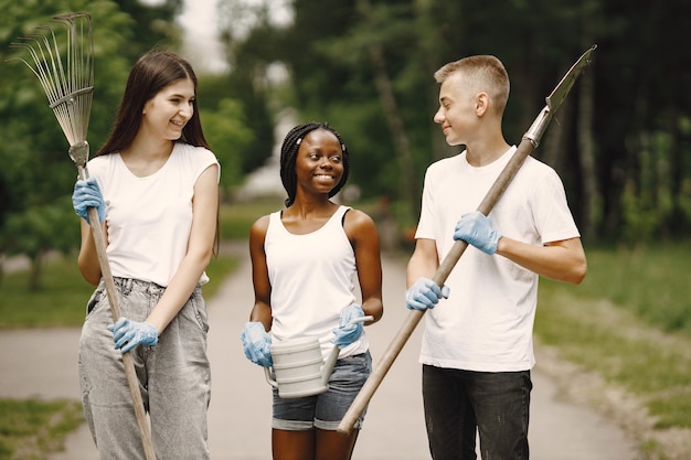 Volunteers working in park. Three teenagers going to plant a tree. Carrying rakes, watering can and seedling. Diverse people.