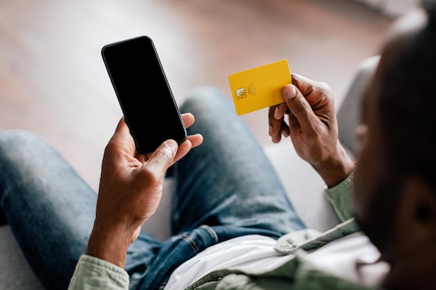 Volwassen Afro-Amerikaanse man typt op smartphone met leeg scherm en toont creditcard in woonkamer