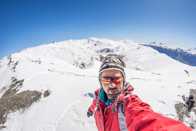 Volwassen alpineskiër met baard, zonnebril en hoed, die selfie op sneeuwhelling in de mooie Italiaanse Alpen met duidelijke blauwe hemel nemen. Concept reislust en avonturen op de berg