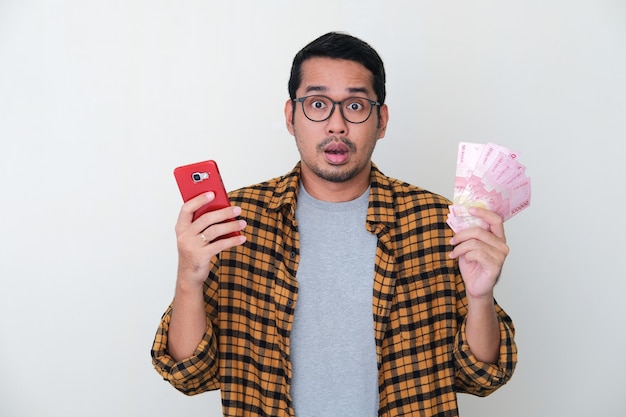 Volwassen Aziatische man die een verwarde gezichtsuitdrukking laat zien terwijl hij een mobiele telefoon en papiergeld vasthoudt