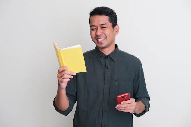 Volwassen Aziatische man die lacht, ontspant terwijl hij een boek leest en een mobiele telefoon vasthoudt