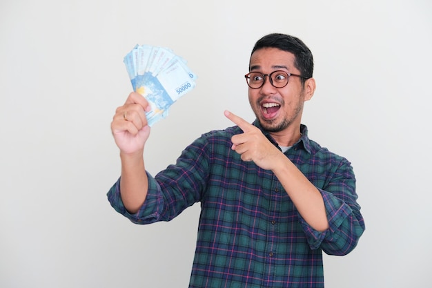 Volwassen Aziatische man die wow-expressie toont terwijl hij naar het geld wijst dat hij vasthoudt