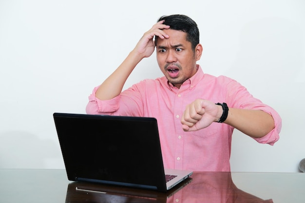 Volwassen aziatische man kijkt naar zijn laptop met een bezorgde uitdrukking terwijl hij zijn horloge laat zien