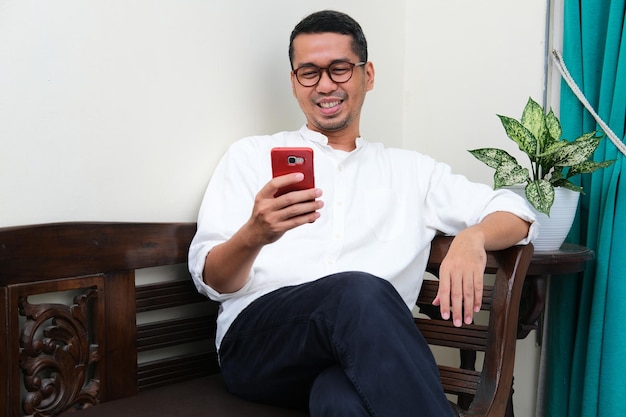 Volwassen Aziatische man zittend op een bank glimlachend wanneer hij naar zijn gsm kijkt