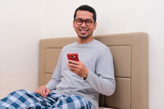 Volwassen Aziatische man zittend op zijn bed glimlachend blij en mobiele telefoon vasthoudend