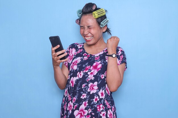 Foto volwassen aziatische vrouw in huisjurk en met haarrollen juichend gelukkig kreeg goed nieuws op smartphone