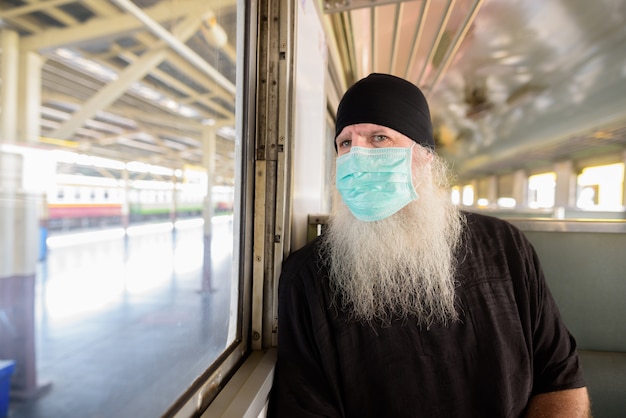 Volwassen bebaarde toeristische man met masker voor bescherming tegen corona virus uitbraak denken in de trein