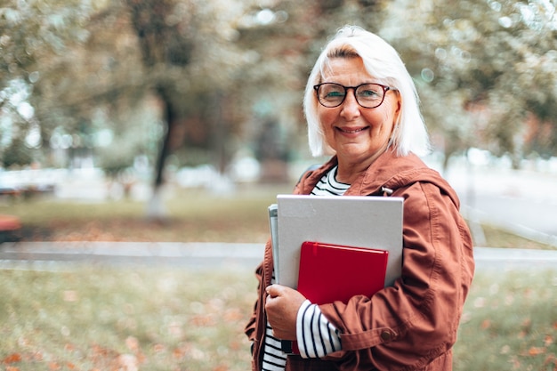 Volwassen blanke blonde vrouw in glazen voor zicht met boeken, notitieboekje en tijdschriften kijken naar de camera wandelen in het herfstpark