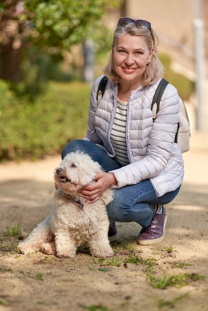 Volwassen blonde vrouw die met pluizige witte hond in stadspark loopt.