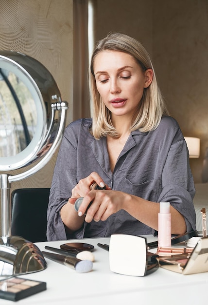 Volwassen blonde vrouw make-up met foundation terwijl ze thuis zit en in de spiegel kijkt