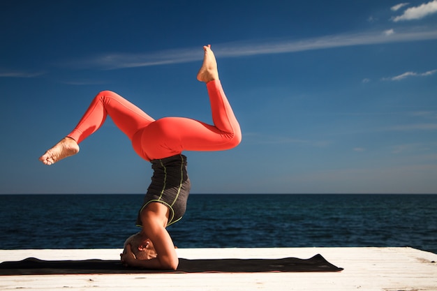 Volwassen blonde vrouw met kort kapsel praktijken yoga op de pier tegen de achtergrond van de zee en de blauwe hemel