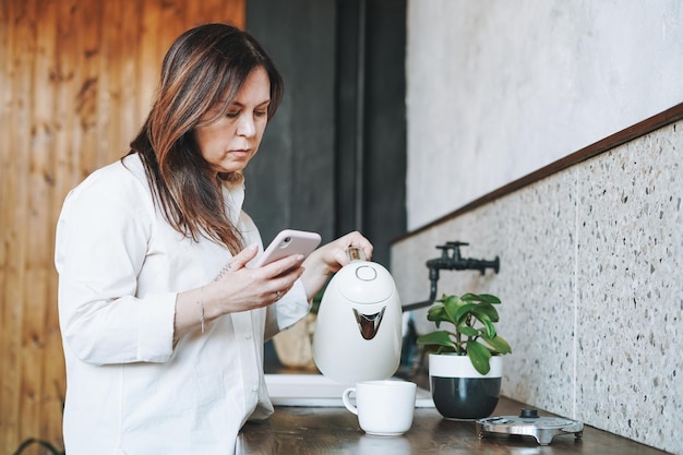 Volwassen brunette lachende vrouw van middelbare leeftijd vijftig jaar in wit overhemd met behulp van mobiele telefoon met kopje koffie in de ochtend in de keuken thuis