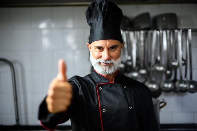 Foto volwassen chef-kok in zwarte jas die duimen omhoog geeft in zijn keuken