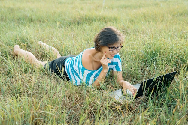 Volwassen glimlachende vrouw die computerlaptop op gras met behulp van