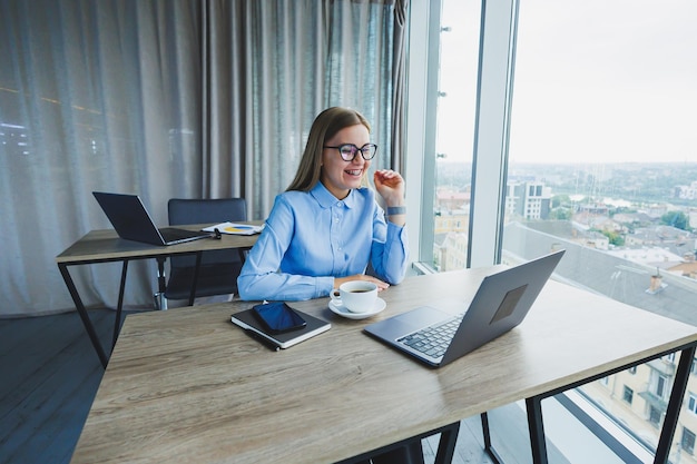 Volwassen glimlachende vrouwelijke manager in bril en formeel shirt zit aan een bureau en kijkt naar een laptop Modern ruim licht kantoor met een groot raam