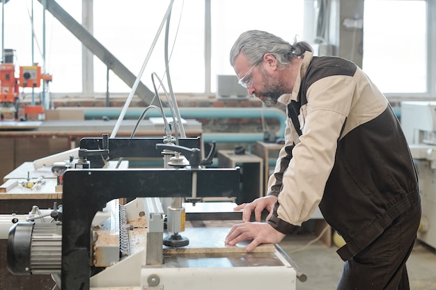 Volwassen ingenieur van meubelfabriek buigen over industriële machine in werkplaats tijdens het bevestigen van werkstuk voor verdere verwerking