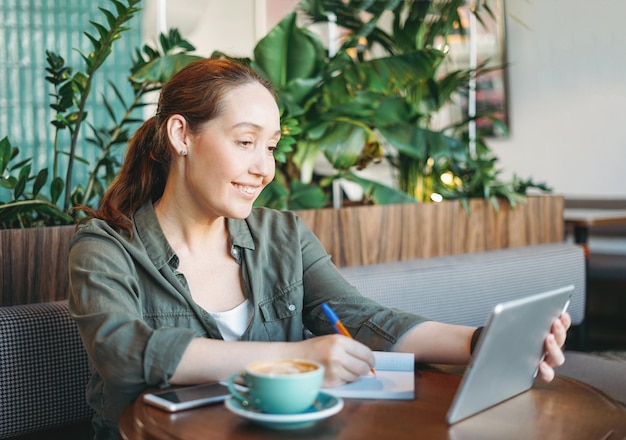 Volwassen lachende brunette vrouw die aantekeningen maakt en naar tablet kijkt, volgt online cursus op tafel in het café