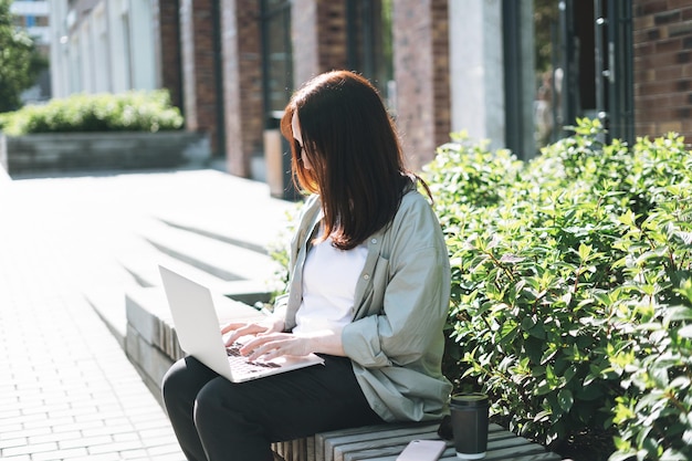 Volwassen lachende brunette zakenvrouw veertig jaar in stijlvol shirt bezig met laptop op openbare plaats op bankje in stadsstraat