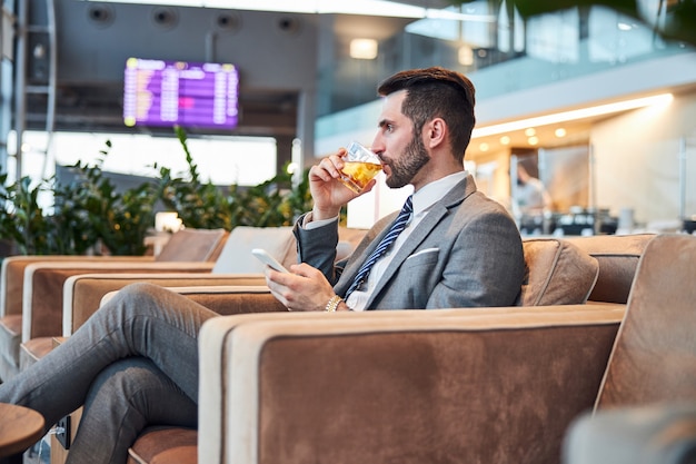 Volwassen man die wat whisky met ijs proeft terwijl hij op de telefoon tikt