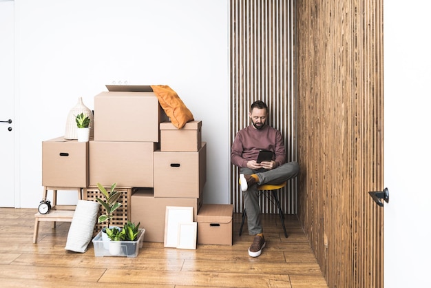Foto volwassen man die zijn tablet gebruikt om dingen uit dozen uit te pakken terwijl hij naar een nieuw ongemeubileerd appartement verhuist