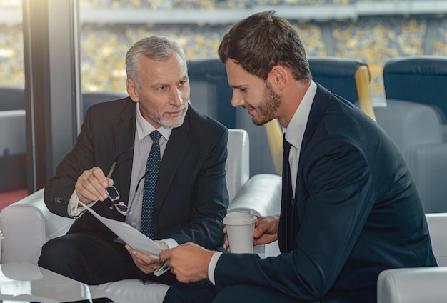 Volwassen man gekleed in pak en zijn werknemer zit in een modern kantoor met documenten in handen en bespreekt iets dat naar papier kijkt