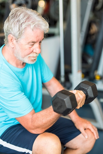 Volwassen man in de sportschool traint zijn lichaam en zijn biceps zittend in een bankje met een halter in zijn hand - gezonde en fitness senior levensstijl