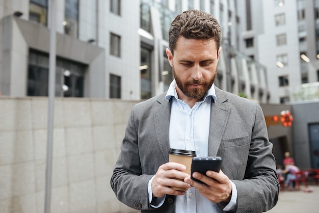 volwassen man in grijs pak mobiele telefoon in de hand kijken, terwijl staan met afhaalmaaltijden koffie voor moderne zakencentrum