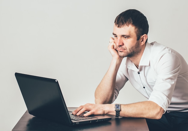 Volwassen man in het witte shirt werkt achter laptop in de kamer. Hij zit en vertrouwt met zijn elleboog op de tafel en onderzoekt de opdracht