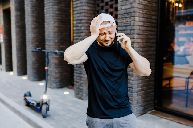 Volwassen man in witte baseballpet en casual kleding die in de buurt van een elektrische scooter staat en op zijn mobiele telefoon op straat praat De man is overstuur en depressief