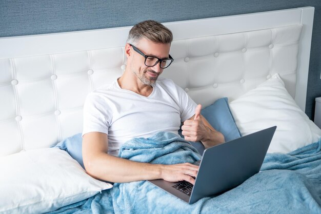 Volwassen man met bril die op laptop werkt in bed videochat