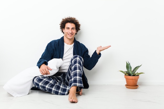 Volwassen man met pyjama zittend op de vloer van het huis met een kopie ruimte op een handpalm en met een andere hand op de taille.