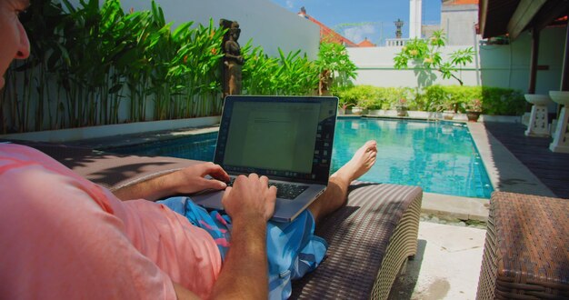 Volwassen man ontspant zich op een leunstoel bij het zwembad en studeert of werkt aan PC-computersoftware Mannelijke handen typen op een laptop toetsenbord terwijl ze buiten rusten achtergrond zomervakantie Remote werken