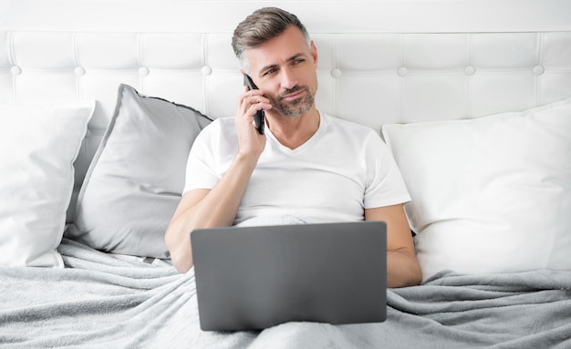 volwassen man spreekt aan de telefoon en werkt op laptop in bed