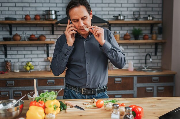 Volwassen man staan aan de keukentafel en praten over de telefoon. Hij proeft een stuk champignon. Man kijk naar beneden. Kleurrijke groenten die op bureau liggen.