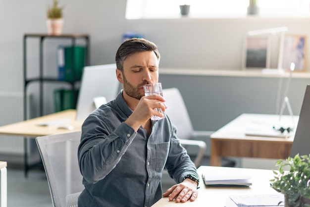 Volwassen mannelijke manager die glas water drinkwater vasthoudt terwijl hij op de computer werkt in een kantoorkopie