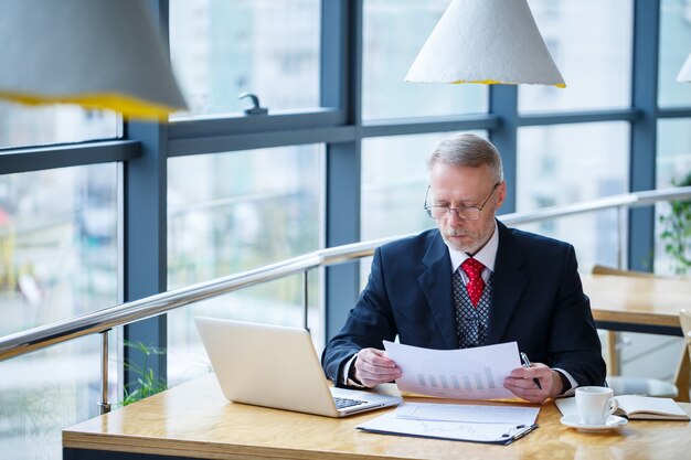 Volwassen mannelijke mentor, regisseur, zakenman in glazen en een pak die documenten bestudeert terwijl hij aan tafel zit. Werkdag concept