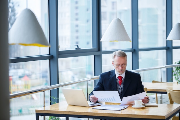 Volwassen mannelijke mentor, regisseur, zakenman in glazen en een pak die documenten bestudeert terwijl hij aan tafel zit. Werkdag concept