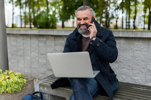 Volwassen mannelijke ondernemer die online werkt op een laptop in een park in een stad die aan de telefoon praat met een