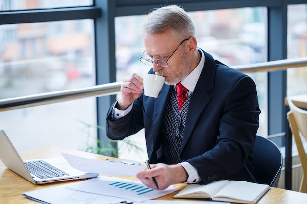 Volwassen mannelijke zakenman die aan een nieuw project werkt en de grafieken van de voorraadgroei bekijkt. Zit aan een groot raam aan de tafel. Kijkt naar het laptopscherm en drinkt koffie.