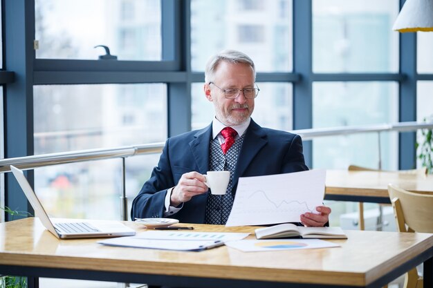 Volwassen mannelijke zakenman die aan een nieuw project werkt en de grafieken van de voorraadgroei bekijkt. Zit aan een groot raam aan de tafel. Kijkt naar het laptopscherm en drinkt koffie.