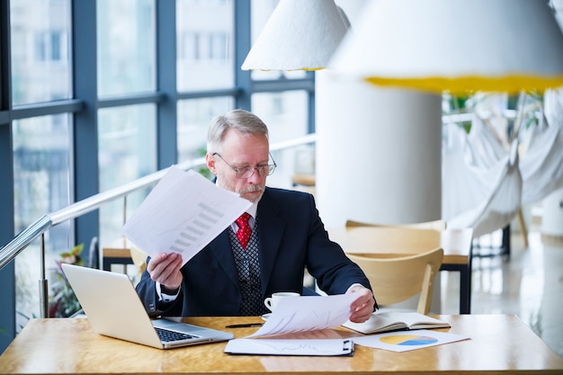 Volwassen mannelijke zakenman die aan een nieuw project werkt en de grafieken van de voorraadgroei bekijkt. zit aan een groot raam aan de tafel. kijkt naar het laptopscherm en drinkt koffie.