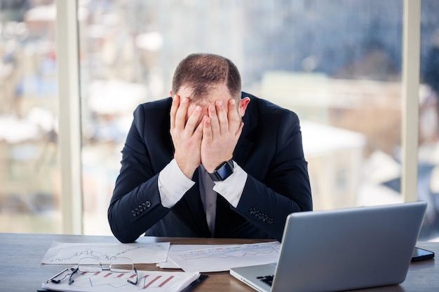 Volwassen mannelijke zakenman, leraar, mentor werkt aan een nieuw project op kantoor en is nerveus. Zit aan de tafel bij het grote raam. Hij werkt op een laptop, zakelijke problemen